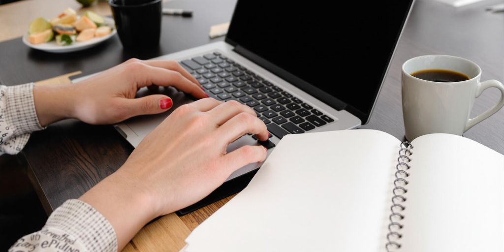 Hands typing on laptop with notebook and coffee nearby.