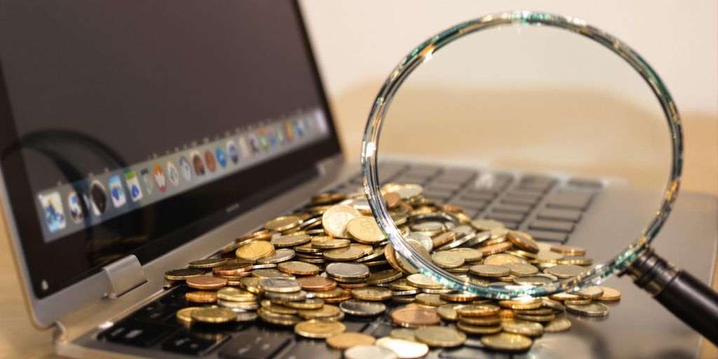 Laptop with coins spilling out, magnifying glass